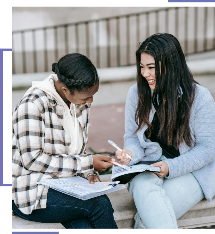 Two women sitting on a bench and one is writing in her notebook.